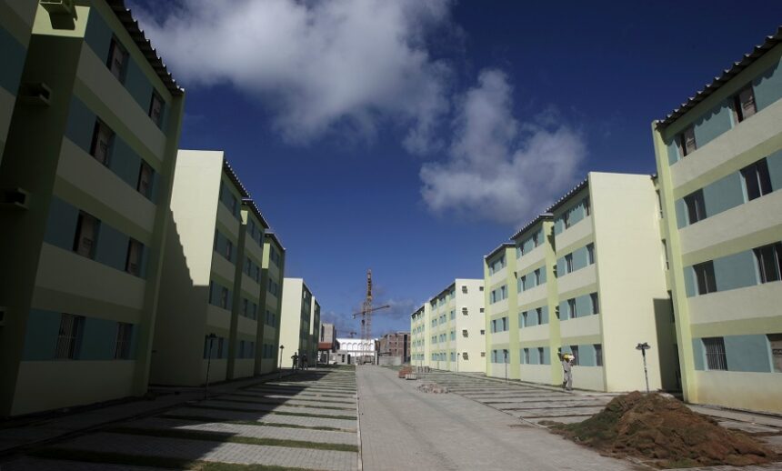 Conjunto habitacional com prédios residenciais em tons de verde, céu azul, e uma grua ao fundo.