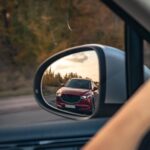 Motorista observa carro vermelho no espelho retrovisor, refletindo cenário de estrada e árvores ao entardecer.