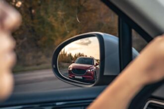 Motorista observa carro vermelho no espelho retrovisor, refletindo cenário de estrada e árvores ao entardecer.
