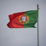 Bandeira de Portugal tremulando ao vento, destacando as cores verde e vermelha e o escudo central em fundo nublado.