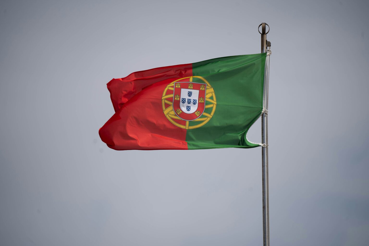 Bandeira de Portugal tremulando ao vento, destacando as cores verde e vermelha e o escudo central em fundo nublado.