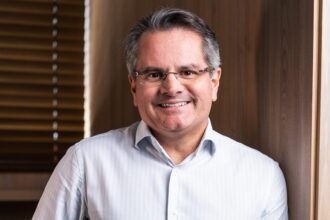 Pessoa sorrindo com óculos e camisa social branca, em um ambiente interno com parede de madeira ao fundo, destacando um clima profissional.