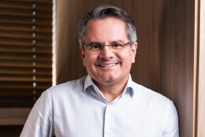 Pessoa sorrindo com óculos e camisa social branca, em um ambiente interno com parede de madeira ao fundo, destacando um clima profissional.