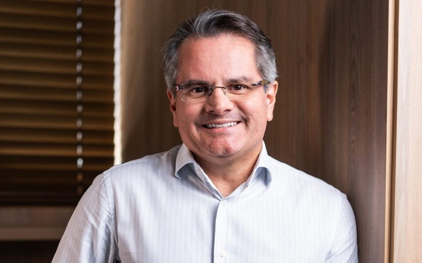Pessoa sorrindo com óculos e camisa social branca, em um ambiente interno com parede de madeira ao fundo, destacando um clima profissional.