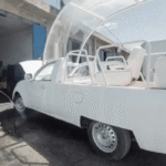Homem lavando veículo modificado com jato d'água em oficina; carro adaptado com assentos e estrutura transparente.