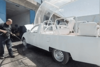 Homem lavando veículo modificado com jato d'água em oficina; carro adaptado com assentos e estrutura transparente.