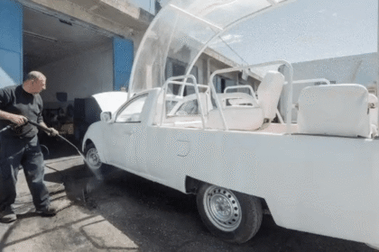 Homem lavando veículo modificado com jato d'água em oficina; carro adaptado com assentos e estrutura transparente.