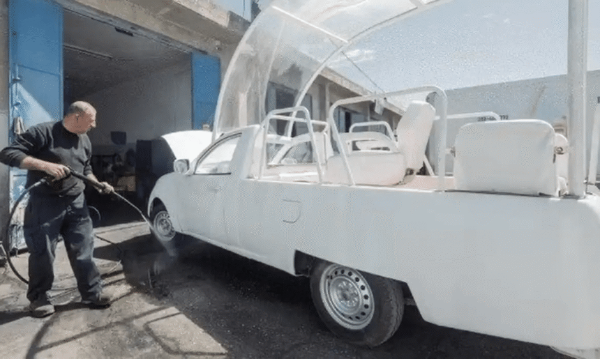 Homem lavando veículo modificado com jato d'água em oficina; carro adaptado com assentos e estrutura transparente.