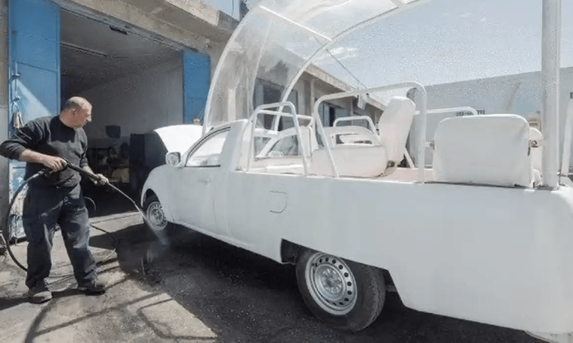 Homem lavando veículo modificado com jato d'água em oficina; carro adaptado com assentos e estrutura transparente.
