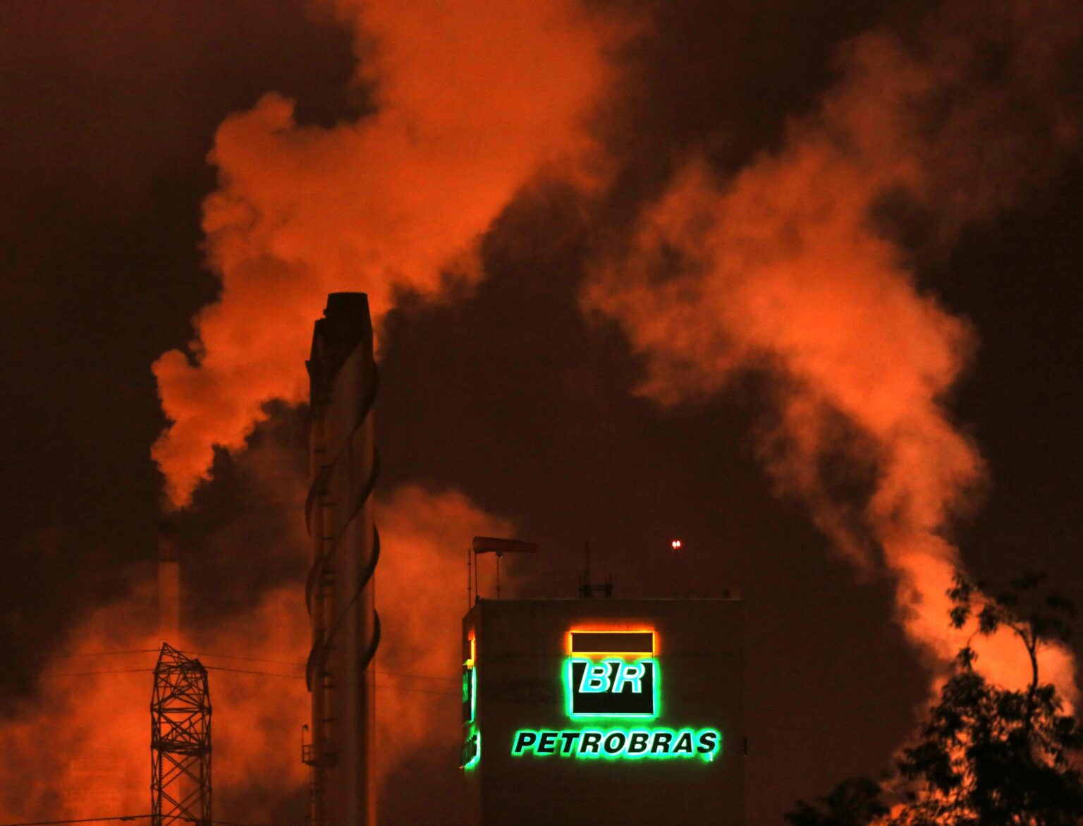 Fumaça laranja sobe de uma refinaria da Petrobras à noite, destacando o logotipo iluminado em meio à poluição industrial.