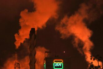 Fumaça laranja sobe de uma refinaria da Petrobras à noite, destacando o logotipo iluminado em meio à poluição industrial.
