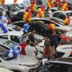 Feira de carros com várias pessoas examinando veículos decorados com balões coloridos, destacando oferta e negociação.