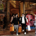 Clientes saem de loja de luxo segurando sacolas de compras; manequins coloridos adornam a vitrine ao fundo.