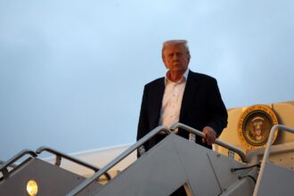 Homem descendo escada de avião presidencial, com o selo dos Estados Unidos ao fundo, sob céu azul claro.