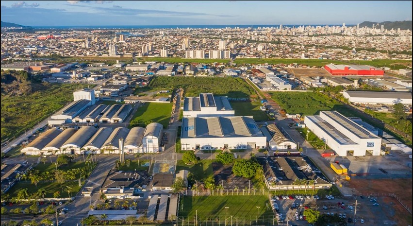 Vista aérea de um complexo industrial em área urbana, com galpões, prédios e vegetação ao redor, cidade ao fundo.