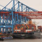 Navio de carga no Terminal de Contêineres de Altenwerder, cercado por guindastes azuis e vermelhos, movimentando contêineres coloridos.
