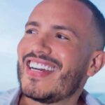 Um homem sorridente segurando uma xícara de café. Ele tem cabelo curto e barba bem aparada. O fundo mostra uma vista para o mar, sugerindo um ambiente relaxante. O homem está usando uma camisa branca aberta, revelando parte do peito.