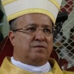 Bispo sentado usando mitra branca com detalhes dourados e vestes litúrgicas douradas. Fundo desfocado com pessoas e grade metálica.