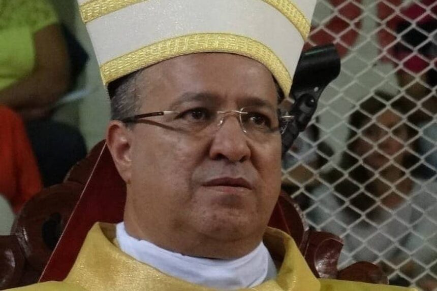 Bispo sentado usando mitra branca com detalhes dourados e vestes litúrgicas douradas. Fundo desfocado com pessoas e grade metálica.