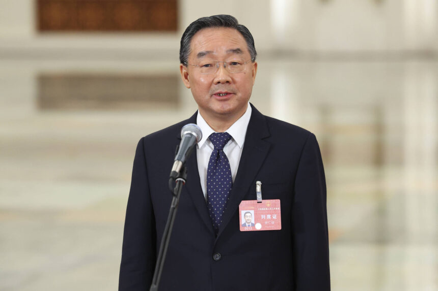 Homem de meia-idade veste terno preto, camisa branca e gravata azul, falando em microfone com crachá preso ao paletó. Ambiente interno amplo com piso de mármore e paredes claras ao fundo.