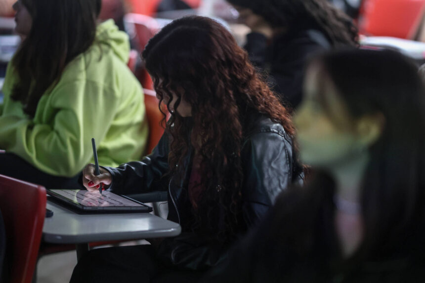 Redação Enem: dicas para escrever sobre IA e era digital A imagem mostra uma estudante sentada em uma sala de aula, concentrada em um tablet que está apoiado em uma mesa. Ela está usando uma caneta stylus para interagir com a tela. A estudante tem cabelos cacheados e está vestindo uma jaqueta preta. Ao fundo, outras pessoas estão visíveis, algumas com roupas de cores variadas, como verde e preto, e a iluminação é suave.