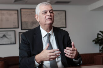 Um homem de terno preto e gravata cinza está sentado em uma mesa, gesticulando com as mãos enquanto fala. Ele tem cabelo grisalho e uma expressão séria. Ao fundo, há quadros com desenhos ou gráficos nas paredes e uma planta em um canto. O ambiente parece ser um escritório moderno.