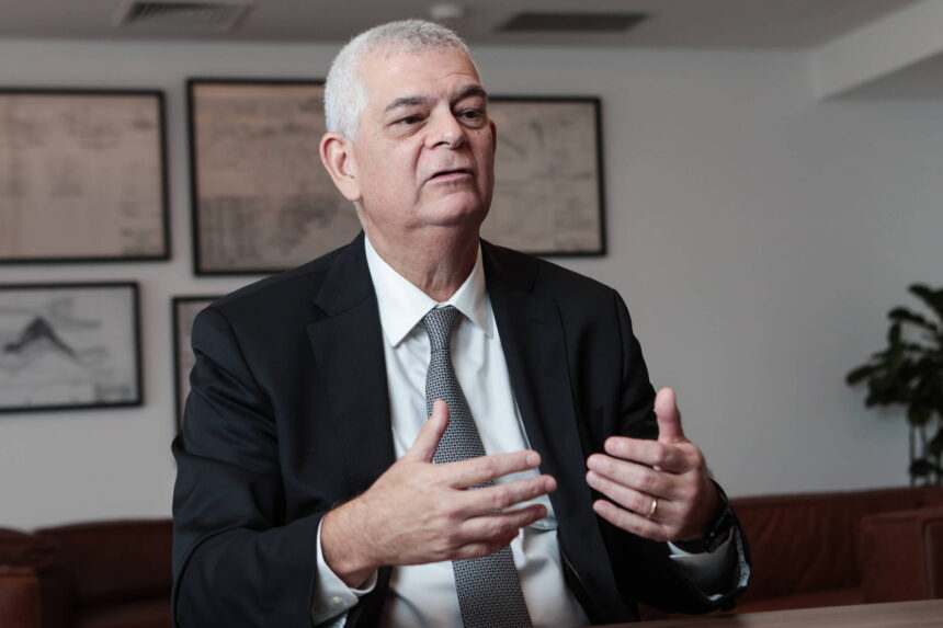 Eletrobras muda nome para Axia Energia em 22 de outubro Um homem de terno preto e gravata cinza está sentado em uma mesa, gesticulando com as mãos enquanto fala. Ele tem cabelo grisalho e uma expressão séria. Ao fundo, há quadros com desenhos ou gráficos nas paredes e uma planta em um canto. O ambiente parece ser um escritório moderno.