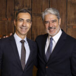 Globo reorganiza telejornais com nova grade de programação A imagem mostra dois homens sorrindo, posando juntos. O homem à esquerda está usando um terno escuro com uma gravata listrada e uma camisa branca. O homem à direita está vestido com um terno escuro e uma gravata com padrão. Ambos têm cabelos curtos e estão em um ambiente com fundo de madeira. Eles parecem estar em um evento formal.