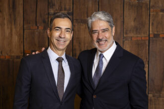 A imagem mostra dois homens sorrindo, posando juntos. O homem à esquerda está usando um terno escuro com uma gravata listrada e uma camisa branca. O homem à direita está vestido com um terno escuro e uma gravata com padrão. Ambos têm cabelos curtos e estão em um ambiente com fundo de madeira. Eles parecem estar em um evento formal.