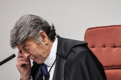 Um juiz está sentado em uma cadeira, com a cabeça inclinada e a mão na testa, parecendo pensativo ou preocupado. Ele usa uma toga preta e uma camisa branca com uma gravata azul. O fundo é neutro, com uma parede clara e uma cadeira vermelha ao fundo.