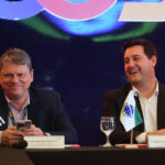 Tarcísio e Ratinho Jr. traçam estratégias para crise no RJ Dois homens sentados lado a lado em mesa de conferência, cada um com uma taça de água à frente. O homem à esquerda segura um celular e tem a bandeira do estado de São Paulo ao seu lado. O homem à direita sorri e tem a bandeira do Brasil à sua frente. Ambos vestem paletó escuro e camisa clara, com fundo escuro e iluminação azulada.