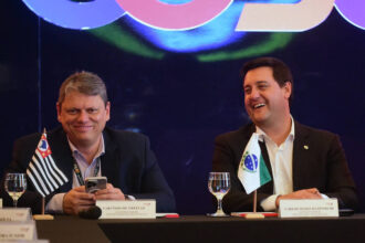 Dois homens sentados lado a lado em mesa de conferência, cada um com uma taça de água à frente. O homem à esquerda segura um celular e tem a bandeira do estado de São Paulo ao seu lado. O homem à direita sorri e tem a bandeira do Brasil à sua frente. Ambos vestem paletó escuro e camisa clara, com fundo escuro e iluminação azulada.