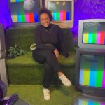 Pessoa sentada em sofá verde cercada por cinco televisores antigos exibindo tela de teste colorida. Ambiente iluminado com luz roxa e decoração com folhas na parede.