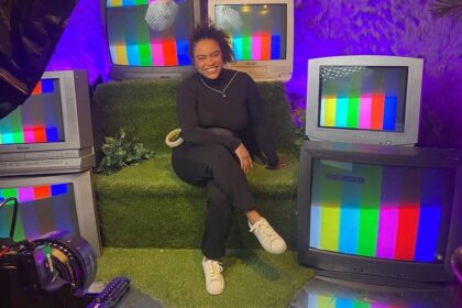 Pessoa sentada em sofá verde cercada por cinco televisores antigos exibindo tela de teste colorida. Ambiente iluminado com luz roxa e decoração com folhas na parede.