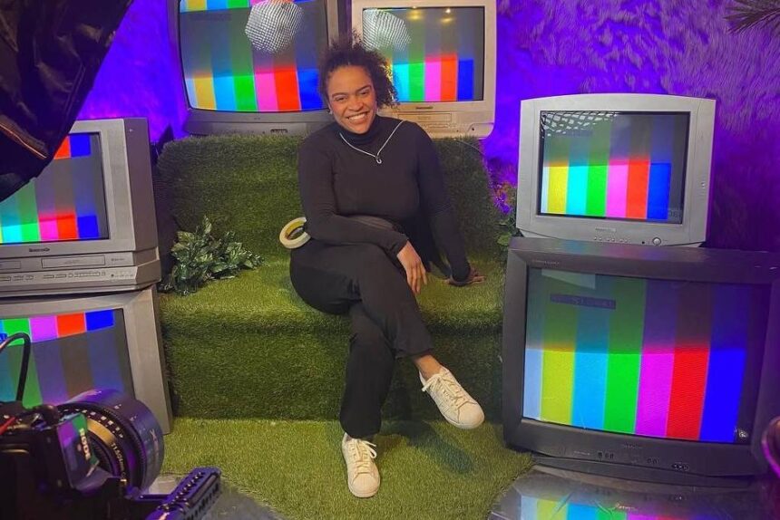 Pessoa sentada em sofá verde cercada por cinco televisores antigos exibindo tela de teste colorida. Ambiente iluminado com luz roxa e decoração com folhas na parede.