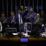 Dez homens vestidos com ternos escuros estão reunidos em plenário. Um homem ao centro está sentado à mesa principal, enquanto os demais ao redor aplaudem e um deles levanta o punho em comemoração. Um cronômetro digital na mesa marca 00:00.