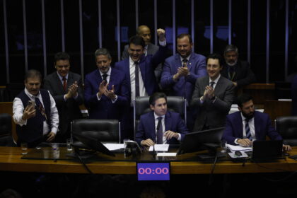 Dez homens vestidos com ternos escuros estão reunidos em plenário. Um homem ao centro está sentado à mesa principal, enquanto os demais ao redor aplaudem e um deles levanta o punho em comemoração. Um cronômetro digital na mesa marca 00:00.