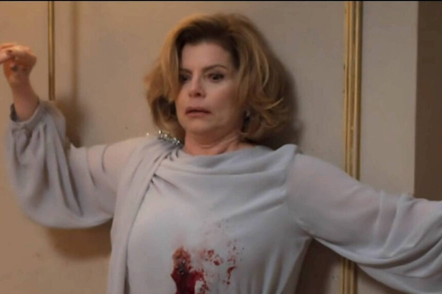 A condenação de Odete Roitman e suas implicações Mulher de cabelos claros e curtos está encostada na parede com os braços levantados. Ela veste blusa cinza clara com mancha vermelha na altura do peito. Ambiente interno com parede bege, molduras douradas, abajur aceso e balde de gelo com garrafa.