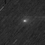 Cometa com núcleo brilhante e cauda difusa visível no centro da imagem, sobre fundo escuro pontilhado por estrelas desfocadas e traços de movimento.
