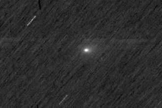 Cometa com núcleo brilhante e cauda difusa visível no centro da imagem, sobre fundo escuro pontilhado por estrelas desfocadas e traços de movimento.