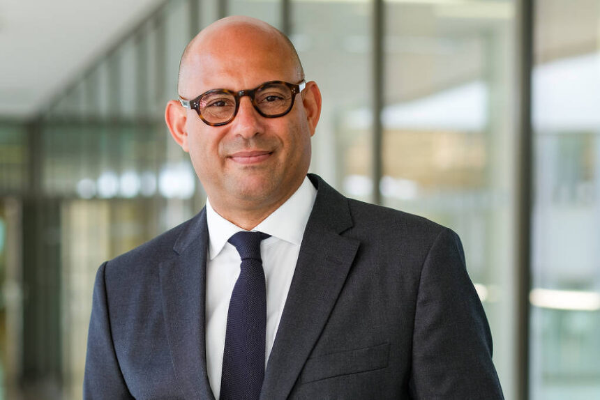Homem calvo com óculos de armação escura veste terno cinza escuro, camisa branca e gravata preta, posando em corredor de escritório moderno com paredes de vidro ao fundo.