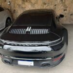 Porsche 911 Turbo S preto estacionado de costas em garagem coberta com piso de cimento. Ao lado esquerdo, há um carro branco estacionado. A parede de fundo é revestida com pedras decorativas. O carro preto tem vidros escurecidos e lanternas traseiras contínuas em LED.