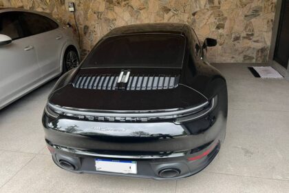 Porsche 911 Turbo S preto estacionado de costas em garagem coberta com piso de cimento. Ao lado esquerdo, há um carro branco estacionado. A parede de fundo é revestida com pedras decorativas. O carro preto tem vidros escurecidos e lanternas traseiras contínuas em LED.