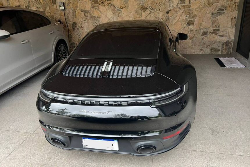 CGU aponta perda em fraude no INSS maior que de 2015 a 2024 Porsche 911 Turbo S preto estacionado de costas em garagem coberta com piso de cimento. Ao lado esquerdo, há um carro branco estacionado. A parede de fundo é revestida com pedras decorativas. O carro preto tem vidros escurecidos e lanternas traseiras contínuas em LED.