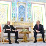 Presidente do Azerbaijão e presidente da Rússia sentados em cadeiras elegantes em sala decorada com padrões geométricos e florais. Bandeiras do Azerbaijão e da Rússia posicionadas ao lado de cada líder, com mesa dourada e arranjo floral entre eles.