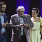 Ruy Castro é reconhecido como autor do melhor livro do ano Quatro pessoas estão alinhadas no palco durante cerimônia. Dois homens no centro seguram troféus, um deles usa blazer cinza e o outro, blazer azul. Homem à esquerda veste terno cinza e gravata azul. Mulher à direita usa vestido amarelo claro com detalhes nos ombros. Fundo escuro com iluminação roxa.