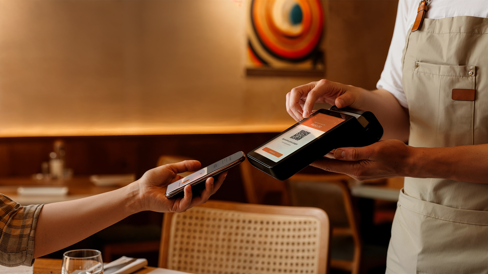 Pessoa faz pagamento por aproximação em restaurante, aproximando o celular de uma maquininha de cartão segurada por um garçom.