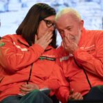 Petrobras tem lucro de R$ 32,7 bilhões no terceiro tri de 2025 A imagem mostra duas pessoas em um evento, vestindo jaquetas laranjas. Elas estão sentadas próximas uma da outra, com expressões de cumplicidade, enquanto uma delas cobre a boca com a mão, como se estivesse sussurrando algo. Ao fundo, há outras pessoas sentadas, mas não estão claramente visíveis. O ambiente parece ser um auditório ou uma sala de conferências.