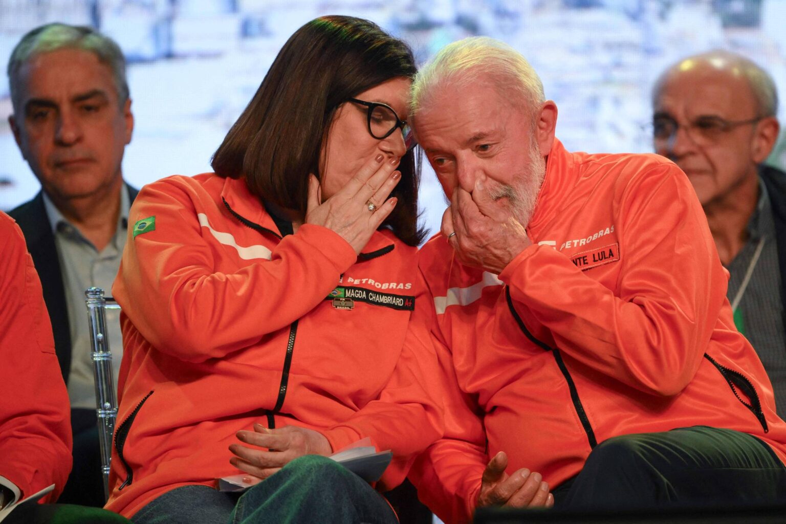 Petrobras tem lucro de R$ 32,7 bilhões no terceiro tri de 2025 A imagem mostra duas pessoas em um evento, vestindo jaquetas laranjas. Elas estão sentadas próximas uma da outra, com expressões de cumplicidade, enquanto uma delas cobre a boca com a mão, como se estivesse sussurrando algo. Ao fundo, há outras pessoas sentadas, mas não estão claramente visíveis. O ambiente parece ser um auditório ou uma sala de conferências.