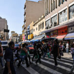 Os nomes e sobrenomes mais comuns no Brasil, segundo o Censo A imagem mostra um cruzamento movimentado em uma área urbana, com várias pessoas atravessando a rua em uma faixa de pedestres. Ao fundo, há prédios de diferentes alturas e estilos arquitetônicos, além de lojas com toldos coloridos. Veículos estão estacionados e em movimento na rua, e o céu está claro, indicando um dia ensolarado.